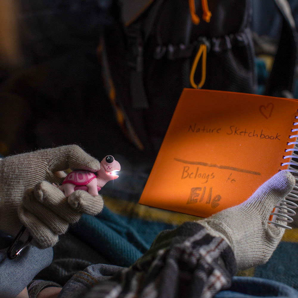 Person holding a small pink creature toy next to an orange nature sketchbook with 'Elle' written on it.